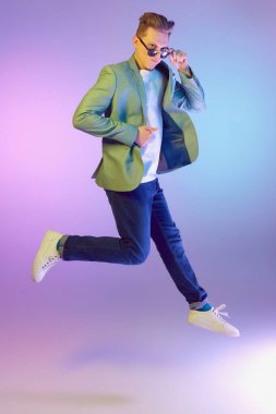 Attentive serious look. Young handsome man in stylish jacket, jeans and sunglasses posing over purple background in neon light. Concept of emotions, lifestyle, youth, modern fashion. Ad