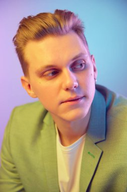 Looking away. Portrait of young handsome man in stylish green jacket posing over gradient blue purple background in neon light. Concept of emotions, lifestyle, youth, modern fashion. Ad