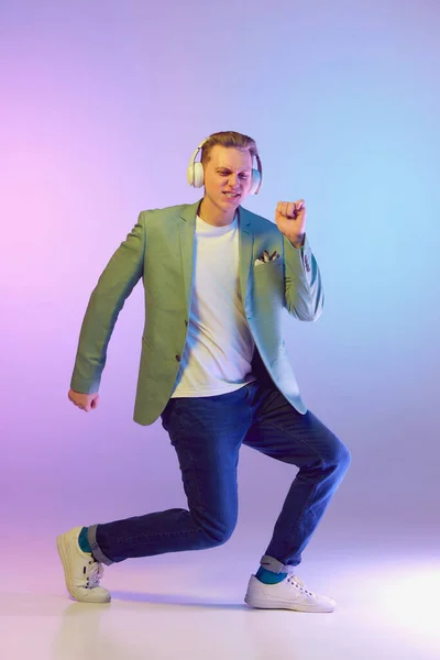 Positive energy. Young handsome man in stylish jacket posing, listening to music in headphones and dancing on purple background in neon light. Concept of emotions, lifestyle, youth, modern fashion. Ad