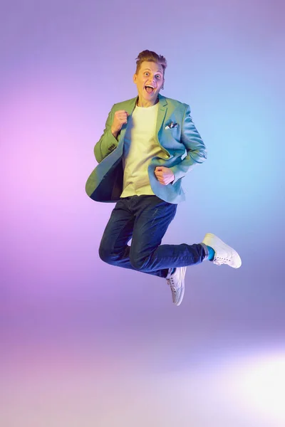Showing emotions of happiness and excitement. Young handsome man in stylish jacket posing in a jump over purple background in neon light. Concept of emotions, lifestyle, youth, modern fashion. Ad