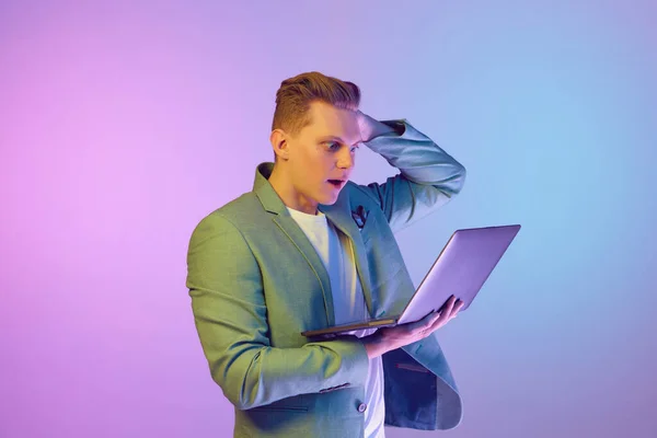 Remote, freelance job. Young handsome man in stylish jacket posing, emotionally looking on laptop over purple background in neon light. Concept of emotions, lifestyle, youth, modern fashion. Ad