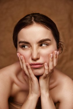 Taking care after sensitive skin with natural cosmetics. Beautiful young brunette girl over brown studio background. Concept of natural beauty, skincare, cosmetology, plastic surgery, health