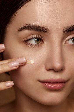 Close-up portrait of beautiful young brunette girl with perfect skin applying cream foundation on face over brown studio background. Concept of natural beauty, skincare, cosmetology, makeup, health