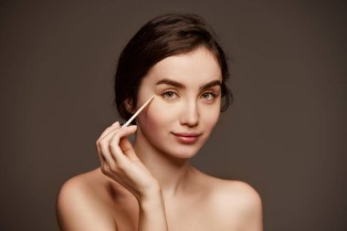 Nude makeup look. Portrait of young beautiful girl with brown hair applying under eye concealer over dark grey studio background. Concept of natural beauty, skincare, cosmetology, cosmetics, health