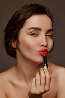 Makeup artist. Portrait of young beautiful girl with brown hair posing with red lipstick over dark grey studio background. Concept of natural beauty, skincare, cosmetology, cosmetics, health