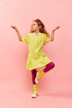 Balancing on one leg. Little cute girl, child with curly hair, colorful hair clips posing in yellow dress over pink studio background. Childhood, emotions, fun, fashion, lifestyle, facial expression