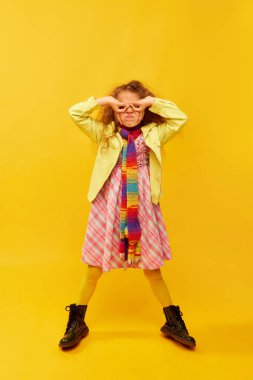Glasses with hands. Little cute girl, child with curly hair emotionally posing in bright clothes on yellow studio background. Concept of childhood, emotions, fun, fashion, lifestyle, facial expression