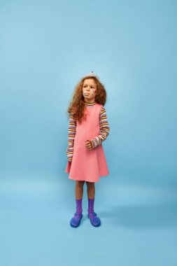 Stickers on face, crown on head. Birthday kid. Little cute girl, child with curly hair posing in pink dress over blue studio background. Childhood, emotions, fun, fashion, lifestyle, facial expression