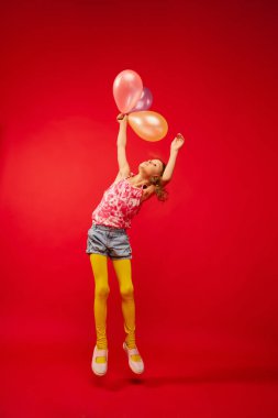 Little cute girl, child with braids emotionally posing, playing, jumping with air balloons on bright red studio background. Concept of childhood, emotions, fun, fashion, lifestyle, facial expression