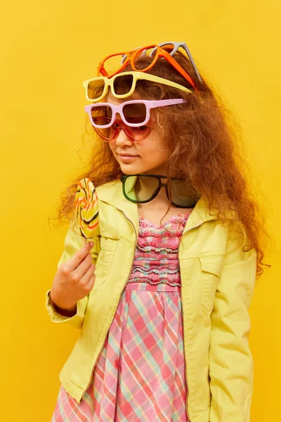 Choosing sunglasses. Little cute girl, child with curly hair posing in bright clothes over yellow studio background. Concept of childhood, emotions, fun, fashion, lifestyle, facial expression