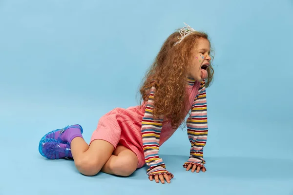 Grimacing, tongue sticking out. Little cute girl, child with curly hair posing in pink dress over blue studio background. Concept of childhood, emotions, fun, fashion, lifestyle, facial expression