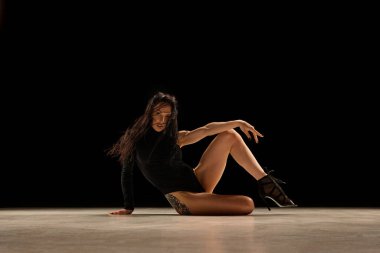 Modern flow. Passionate young woman in bodysuit and heels dancing, performing over black background. Concept of contemporary dance style, art, aesthetics, hobby, creative lifestyle