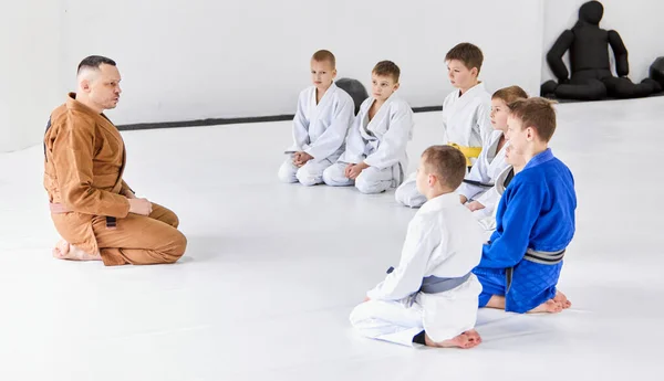 小さな男の子のグループは 子供の着物の床のコーチの周りに座っている 子供たちは屋内で訓練する 柔術の訓練 武道の概念 戦闘スポーツ スポーツ教育 子供の頃 — ストック写真