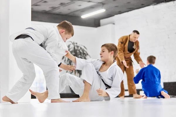 Professional sport activity. Boys, children in white kimono training, practising judo, jiu-jitsu exercises indoors. Concept of martial arts, combat sport, sport education, childhood