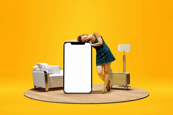 テキストのための空の画面を持つ携帯電話の巨大な3Dモデルにもたれて肯定的な笑顔の若い女の子 自宅のインテリアと黄色の背景の上に広告 フリーランス デザイン ロゴのためのモックアップ — ストック写真