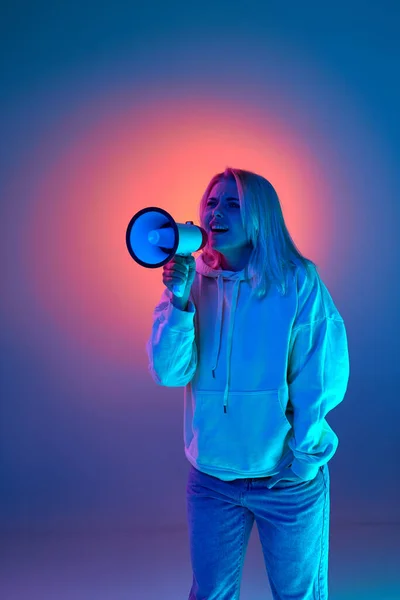 News, information. Young woman talking in megaphone over gradient purple studio background in blue neon light. Concept of emotions, facial expression, lifestyle, inspiration, sales, ad