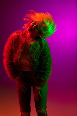 Dancing. Energy. Expressive young woman in fur coat posing over magenta studio background in green neon light. Concept of emotions, fashion, facial expression, lifestyle, inspiration, sales, ad