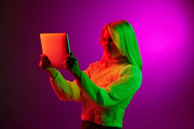 Communication. Emotive young woman having online video call on tablet over magenta studio background in green neon light. Concept of emotions, facial expression, lifestyle, inspiration, sales, ad