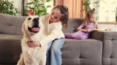 Little girls, children spending time at home in living room. Playing with purebred golden retriever and reading book. Concept of family, childhood, pets, care, friendship, emotions. leisure