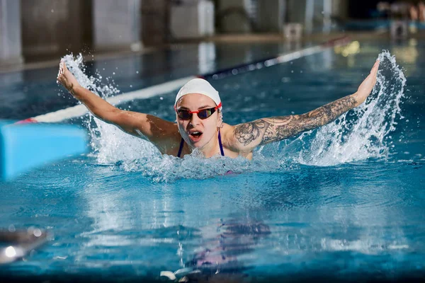 Butterfly swimming type. Young woman, professional female swimming athlete in cap and goggles training in pool indoors. Concept of sport, endurance, competition, energy, healthy lifestyle