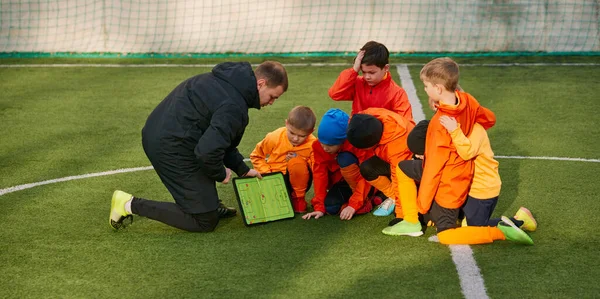 Group of boys, children training football with professional coach on field outdoors. Man showing sportive tactics and positions. Concept of sport, childhood, active lifestyle, hobby, sport club