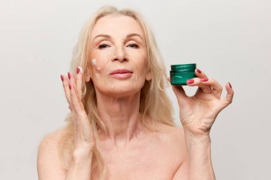 Attractive middle-aged woman applying moisturizing, nourishing face cream. Mature old woman posing against grey background. Concept of natural beauty, face skin care, cosmetology and cosmetics, health