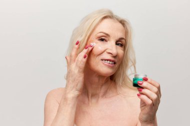 Beautiful middle-aged woman applying moisturizing face cream. Mature old lady posing against grey background. Concept of natural beauty, face skin care, cosmetology and cosmetics, health