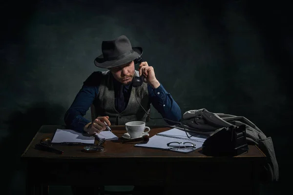 Professional detective in fedora hat sitting at table and talking on phone over dark green vintage background. Investigation. Concept of occupation, character, history. Retro style
