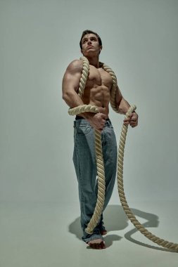 Full-length image of handsome young man with strong muscular body shape posing in jeans with rope over grey studio background. Concept of mans beauty, sportive and healthy lifestyle, fashion