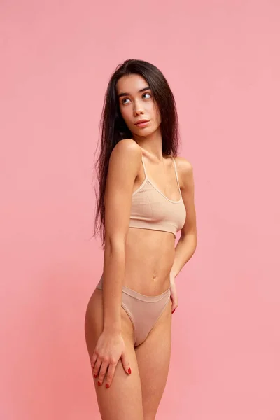 Femininity. Portrait of young tender brunette girl with slim body shape posing in beige underwear over pink studio background. Concept of natural beauty, fitness, diet, body care, sport and health