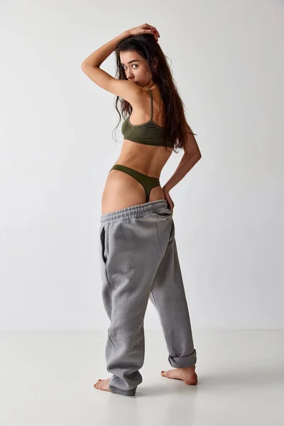 Diet. Portrait of young girl with slim, fit body posing in underwear and oversized pants over grey studio background. Concept of natural beauty, weight loss, body-positivity, wellness, body care