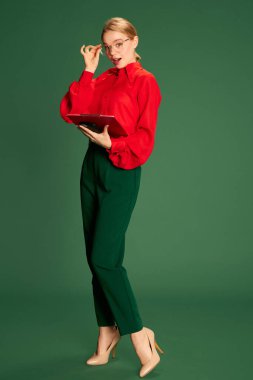 Full-length portrait of young girl, business woman in glasses and red blouse posing over green studio background. Manager. Concept of business, emotions, official fashion, lifestyle
