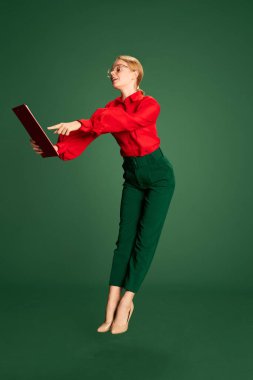 Checking working tasks. Young beautiful girl in stylish clothes looking at documents over green studio background. In a jump. Concept of business, emotions, official fashion, lifestyle