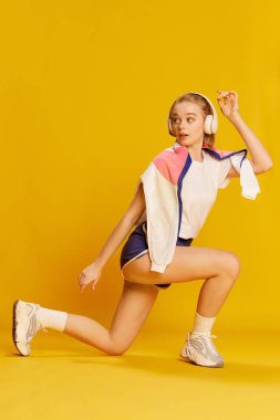 Keeping body fit and healthy. Beautiful young girl in headphones and stylish sportswear training over yellow studio background. Concept of beauty, sportive lifestyle, hobby, emotions. Ad