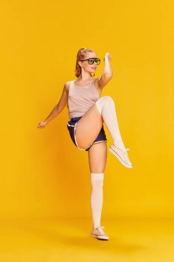 Warming-up exercises. Full-length image of beautiful slim young girl in sunglasses training over yellow studio background. Concept of beauty, sportive lifestyle, hobby, emotions. Ad