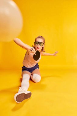 Funny activity. Happy cheerful young girl in sunglasses posing, playing table tennis over yellow studio background. Concept of beauty, sportive lifestyle, fashion, hobby, emotions. Ad