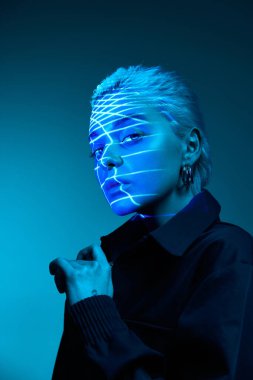 Modern technologies. Portrait of young blonde girl with neon stripes on face posing over dark background in blue neon lights. Concept of art, modern style, cyberpunk, futurism and creativity