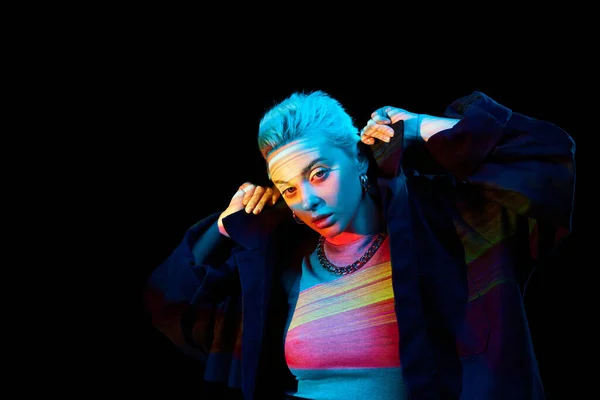 Portrait of young blonde girl with neon multicolored stripes on face posing over dark background in blue neon lights. Concept of art, modern style, cyberpunk, futurism and creativity
