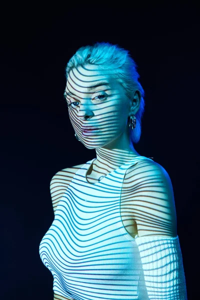 Abstract art. Portrait of young blonde girl with neon stripes on face posing over dark background in blue neon lights. Concept of art, modern style, cyberpunk, futurism and creativity