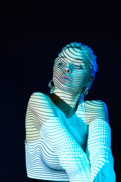 Futuristic. Portrait of young blonde girl with neon stripes on face posing over dark background in blue neon lights. Concept of art, modern style, cyberpunk, futurism and creativity