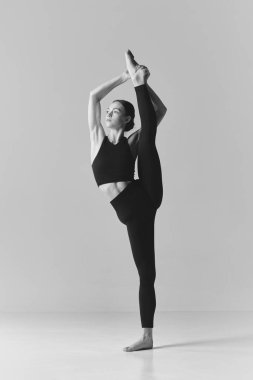 Black and white photography. Young flexible girl doing stretching exercises, standing on twine over studio background. Concept of sport, body care, beauty, fitness, active lifestyle. Ad