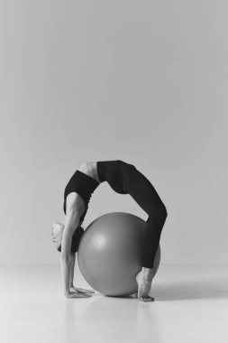 Black and white photography. Young flexible fit girl doing stretching exercises, standing over fitness ball, training over studio background. Concept of sport, body care, fitness, active lifestyle. Ad