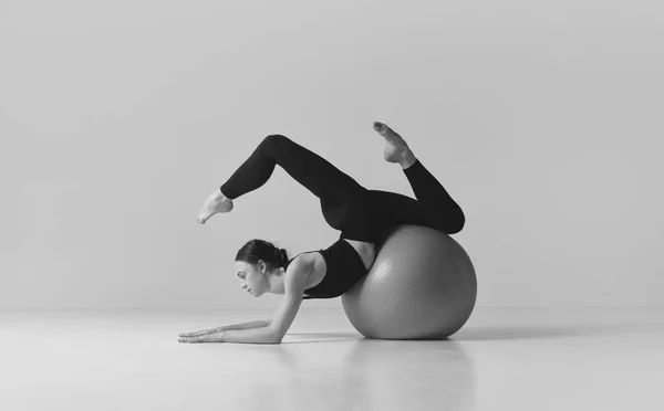 Fitness aesthetics. Black and white photography. Young sportive girl training with fitness ball, balancing over studio background. Concept of sport, body care, beauty, fitness, active lifestyle. Ad