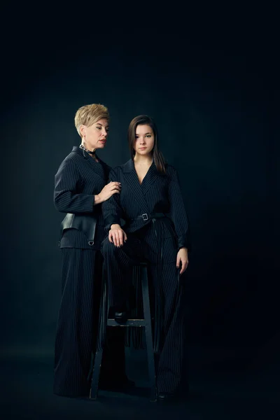 Stylish look. Full-length portrait of middle-aged woman, mother posing with her young daughter over dark studio background. Concept of motherhood, family, mothers day, love, emotions, relationship