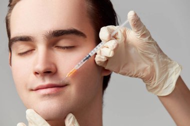 Lip augmentation. Close-up of young man doing beauty cosmetological injections. Male model against grey studio background. Concept of mens health, skin care, hygiene and male cosmetological treatment