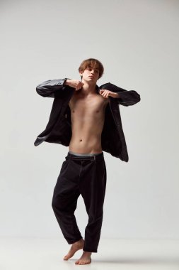 Full-length portrait of young handsome man posing shirtless in leather jacket and black pants against grey studio background. Concept of mens health, modern fashion, beauty, style, body care
