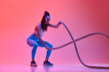 Full-body workout. Fitness. Young fit girl training, doing exercises with rope over pink studio background in neon light. Concept of sportive lifestyle, health, endurance, action and motion. Ad