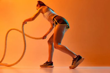 Side view. Workout. Young sportive girl training, doing exercises with rope over orange studio background in neon light. Concept of sportive lifestyle, health, endurance, action and motion. Ad