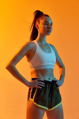 Portrait of young sportive girl with strong muscular body posing over orange studio background in neon light. Concept of sportive lifestyle, health, endurance, action and motion. Ad