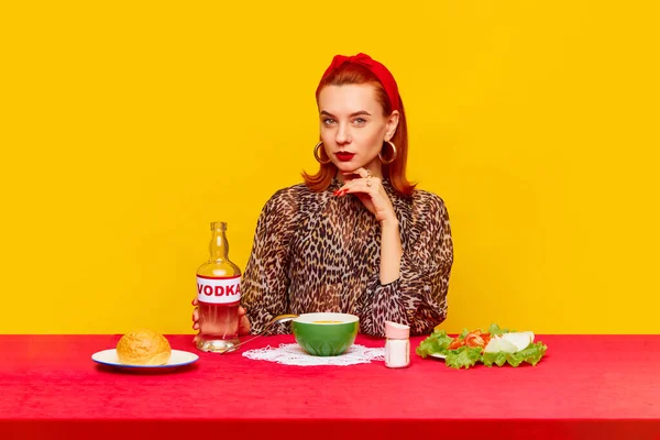 Food pop art photography. Beautiful redhead woman sitting at table with serious face against yellow background and having dinner with vodka and salad. Complementary colors. Copy space for ad, text